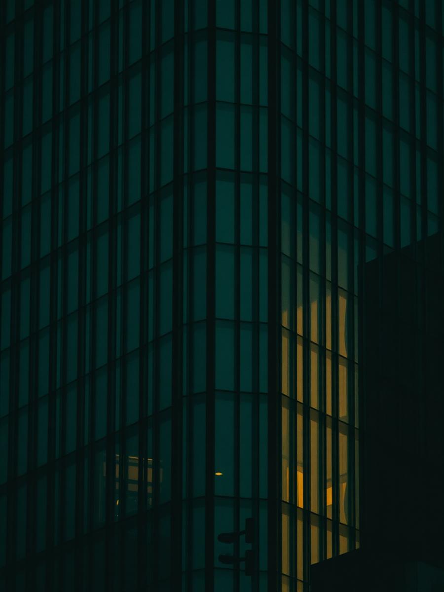 A dark green glass tower at dusk with one window lit gold